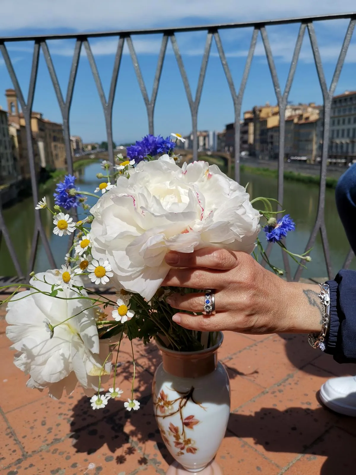 A hand wearing a ring, holding a bunch of peonies and cornflowers on the terrace overlooking the Arno