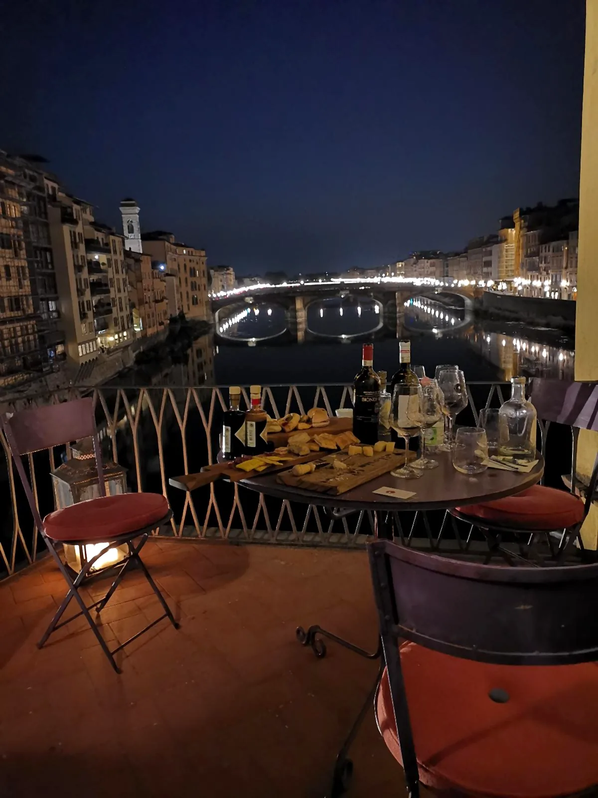 The shop terrace on the Arno, above Ponte Vecchio, in the evening: a small table set for a toast, with Ponte Santa Trinita lit up in the background